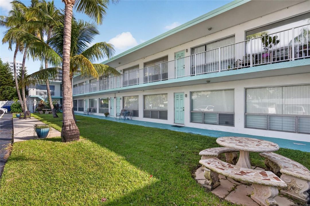 Two-story multi-unit exterior featuring white stucco siding, teal-toned trim, a second-story balcony with metal railings, and a landscaped lawn with palm trees