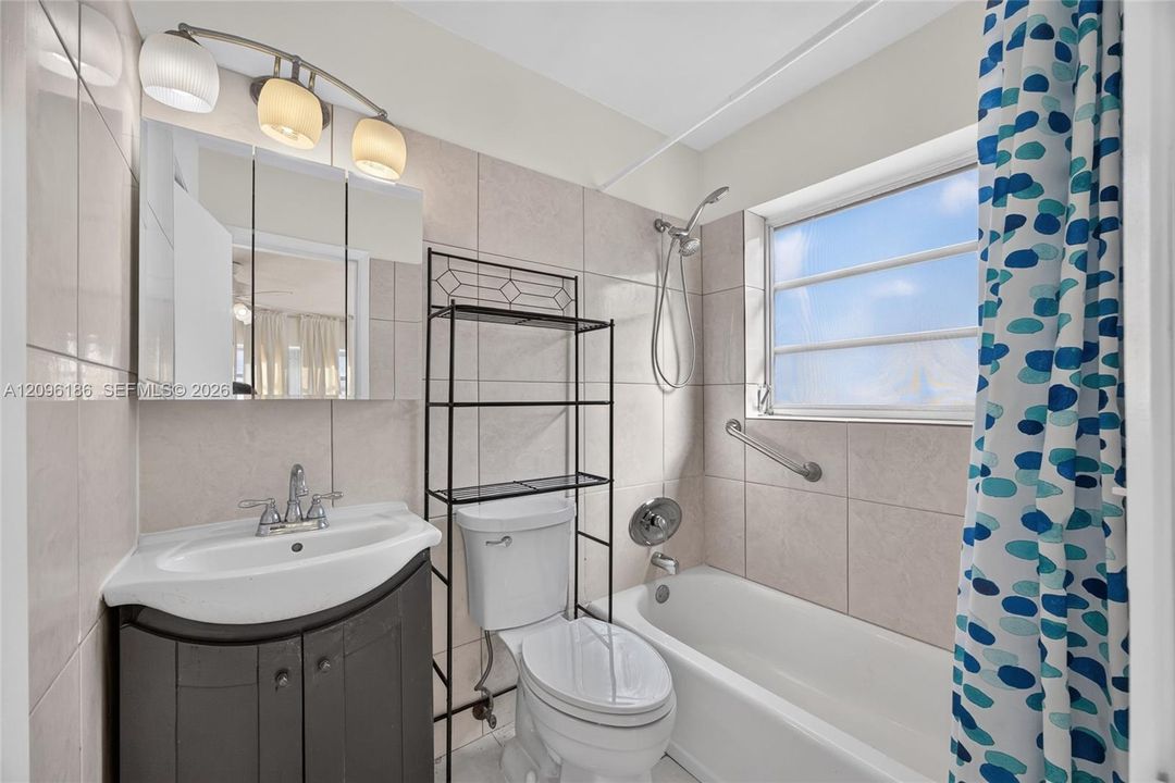 Bathroom featuring a pedestal-style vanity with storage, tile-surround bathtub with a handheld shower head, wall-mounted light fixture, and horizontal-slat window