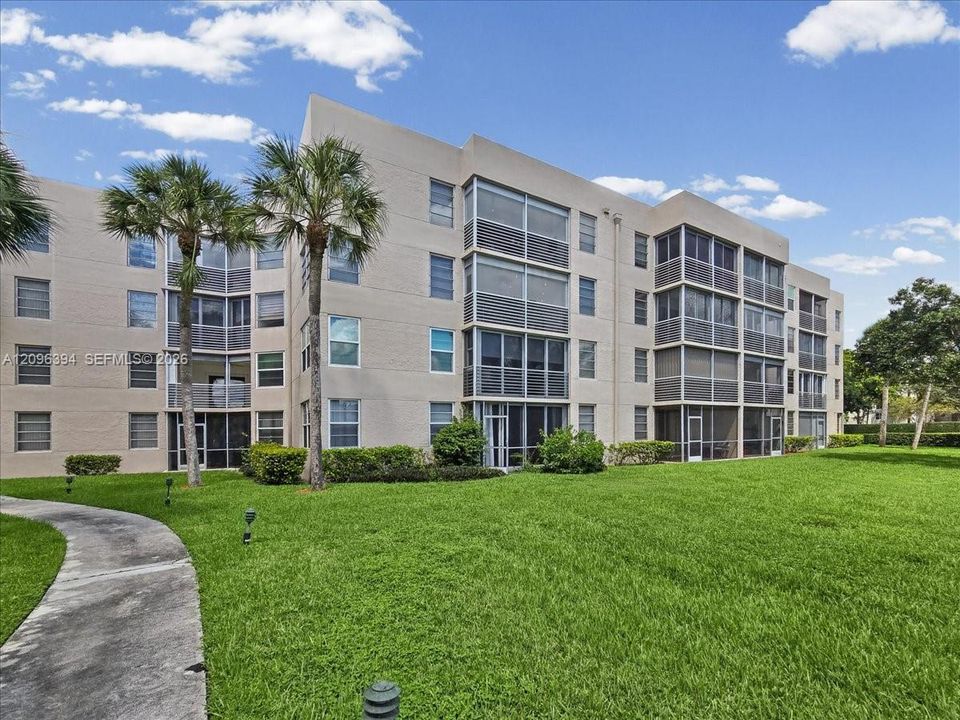 Multi-story residential building featuring a stucco exterior, screened balconies, and a concrete walkway set within a landscaped lawn