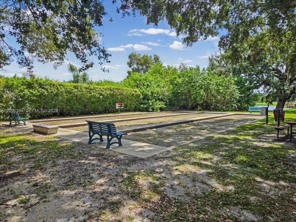 Bocce ball court featuring a rectangular sand playing surface, timber border edging, concrete perimeter walkways, and mature tree-lined surroundings