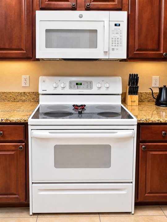 Stained wood cabinetry, white electric range with glass cooktop, built-in white over-the-range microwave, and stone-finish countertops