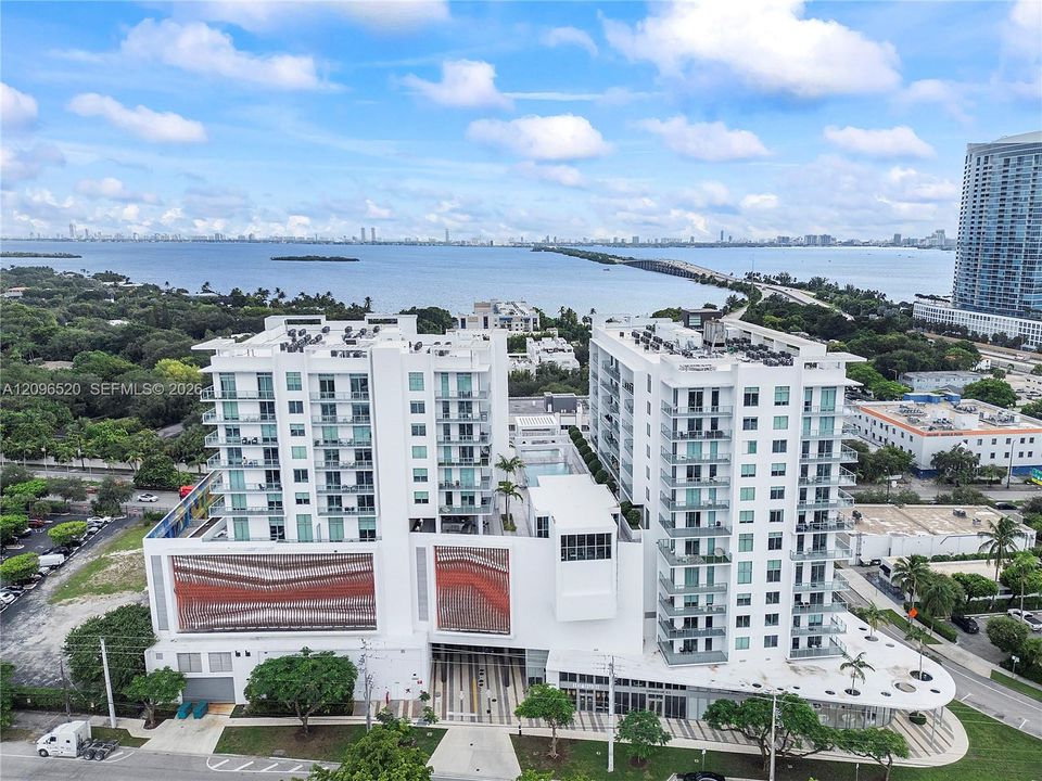 High-rise multi-story exterior with floor-to-ceiling glass balconies, textured facade accents, ground-level covered entryway, and water surroundings