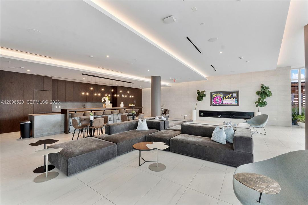 Open-concept lounge featuring large-format tile flooring, recessed cove lighting, a linear built-in fireplace, and dark-toned cabinetry with an integrated bar