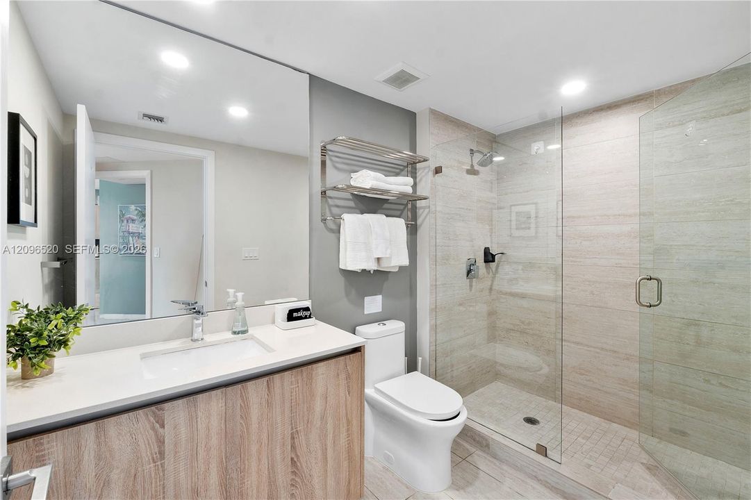 Bathroom featuring a wood-finish vanity with a quartz countertop, integrated sink, frameless glass shower enclosure with horizontal wall tile, and wall-mounted metal shelving
