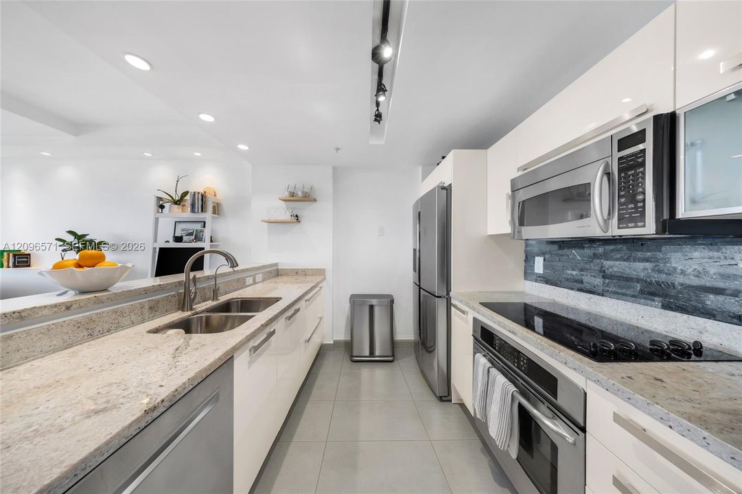 Kitchen featuring high-gloss cabinetry, stone countertops, stainless steel appliances, textured stone backsplash, and recessed track lighting