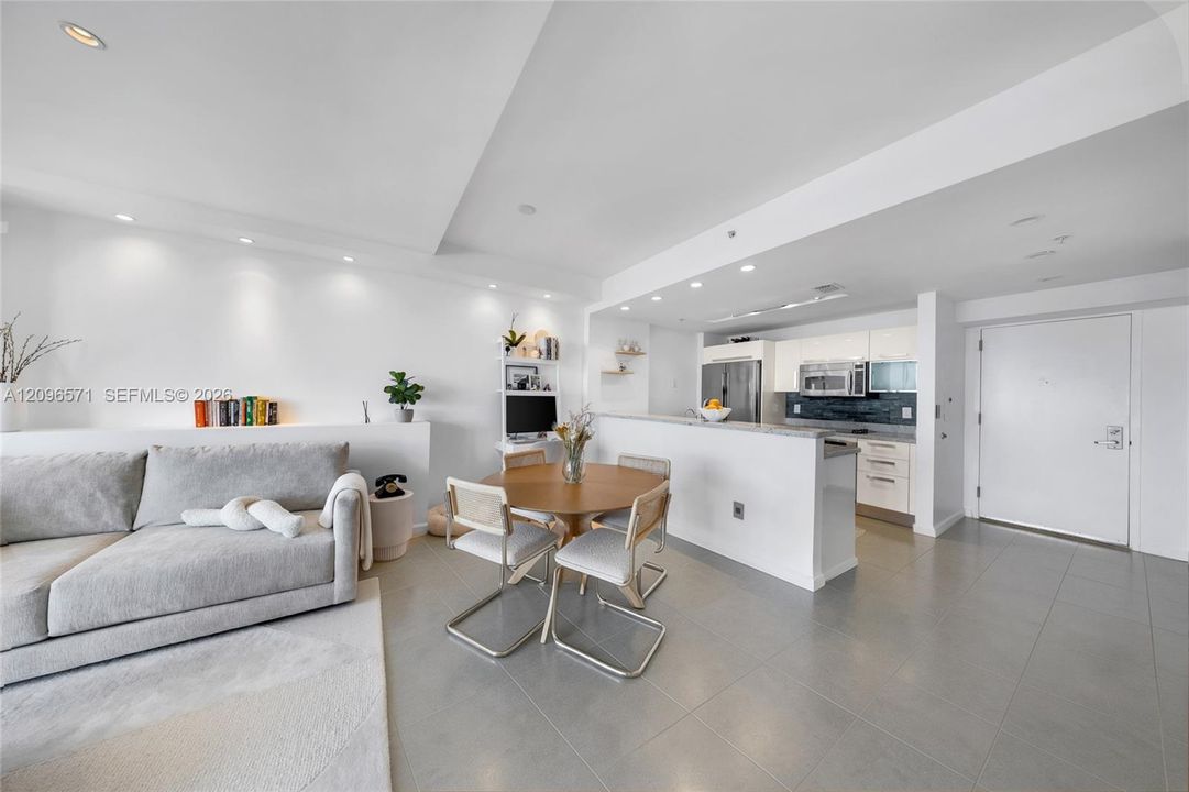 Open-concept interior featuring large-format tile flooring, recessed ceiling lighting, a kitchen peninsula with a stone countertop, white flat-panel cabinetry, and a built-in display niche