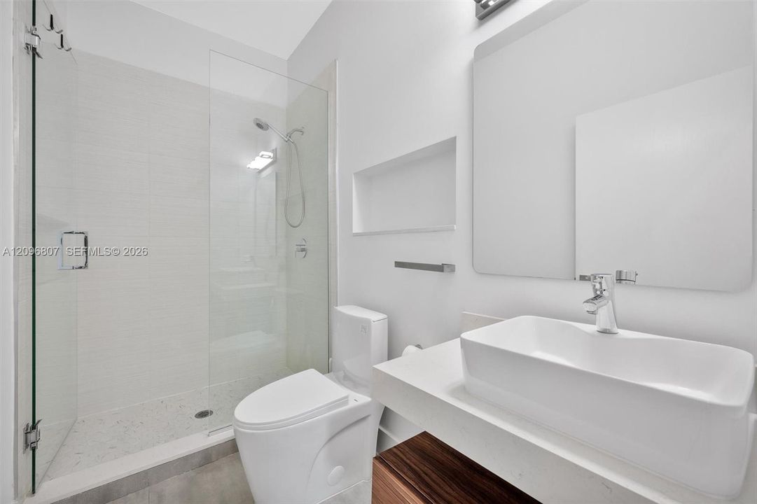 Bathroom featuring a vessel sink with a white stone-composite countertop, wood-finish vanity cabinet, walk-in shower with a glass enclosure and built-in wall niche, and a wall-mounted mirror