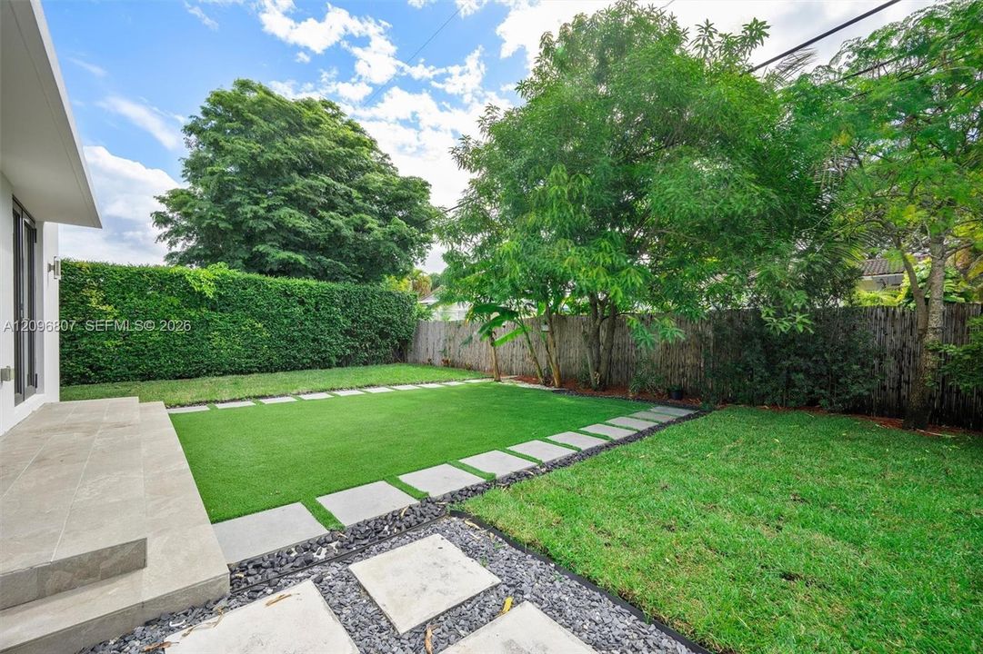 Rear yard featuring a tiled patio, synthetic turf lawn, stone paver pathway, perimeter hedge, and mature trees