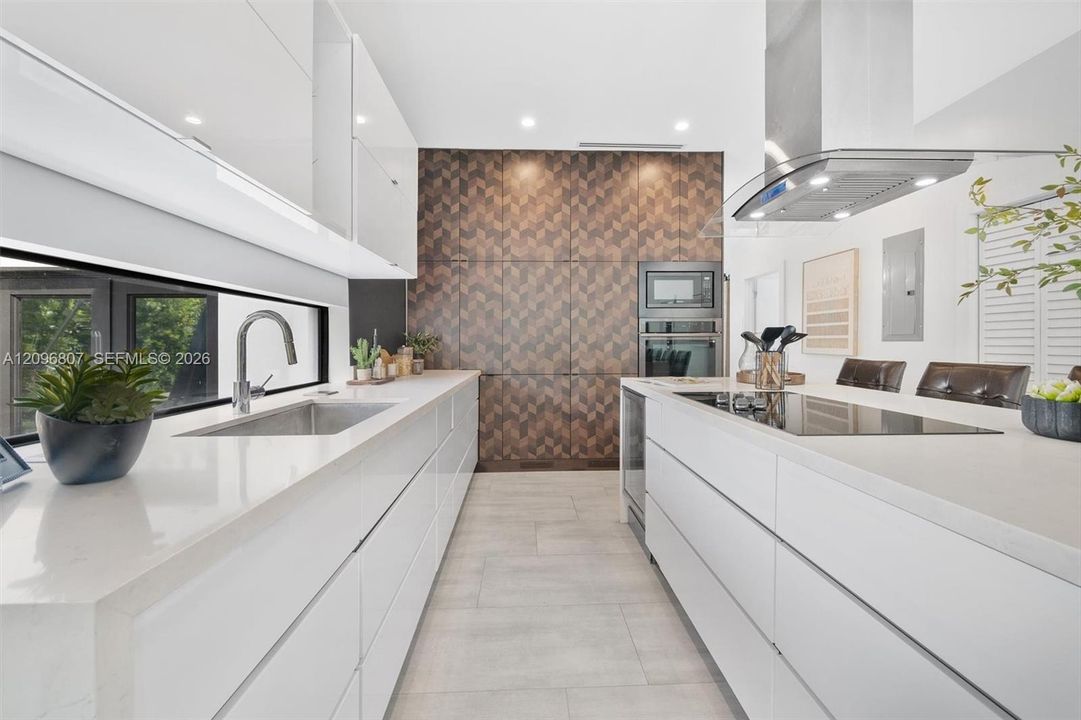Modern kitchen featuring flat-panel white cabinetry, expansive stone countertops, geometric-patterned wall paneling, built-in stainless steel appliances, and a central island with an integrated cooktop