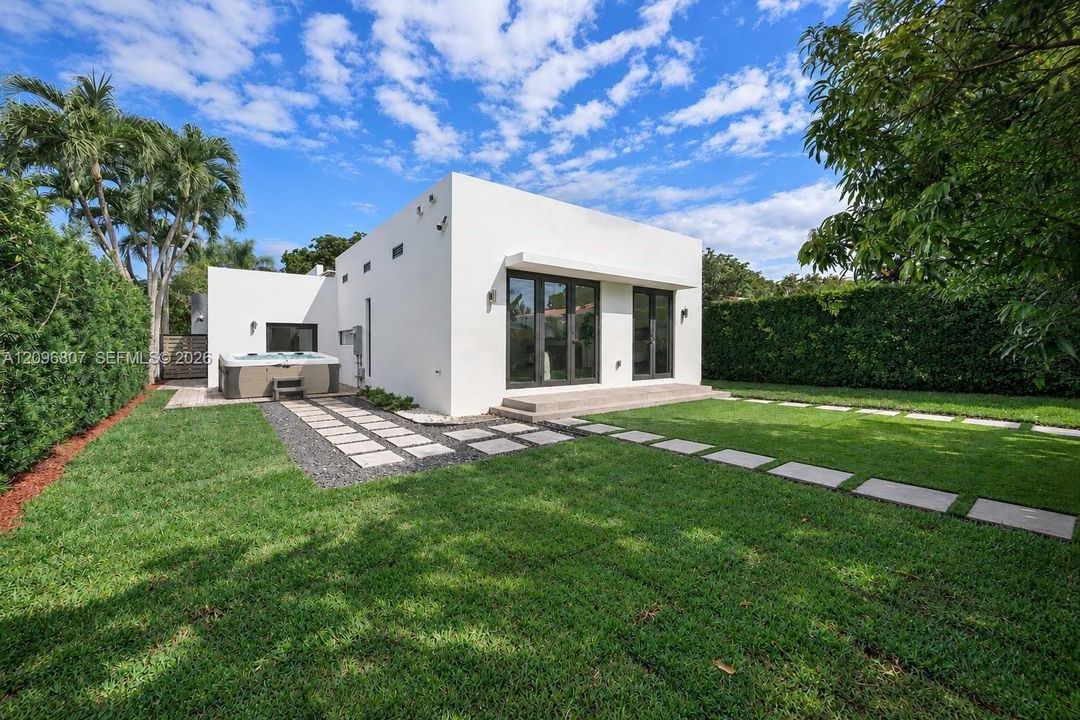 Modern exterior featuring smooth white stucco finish, built-in hot tub, concrete paver pathways, manicured lawn, and perimeter privacy hedges