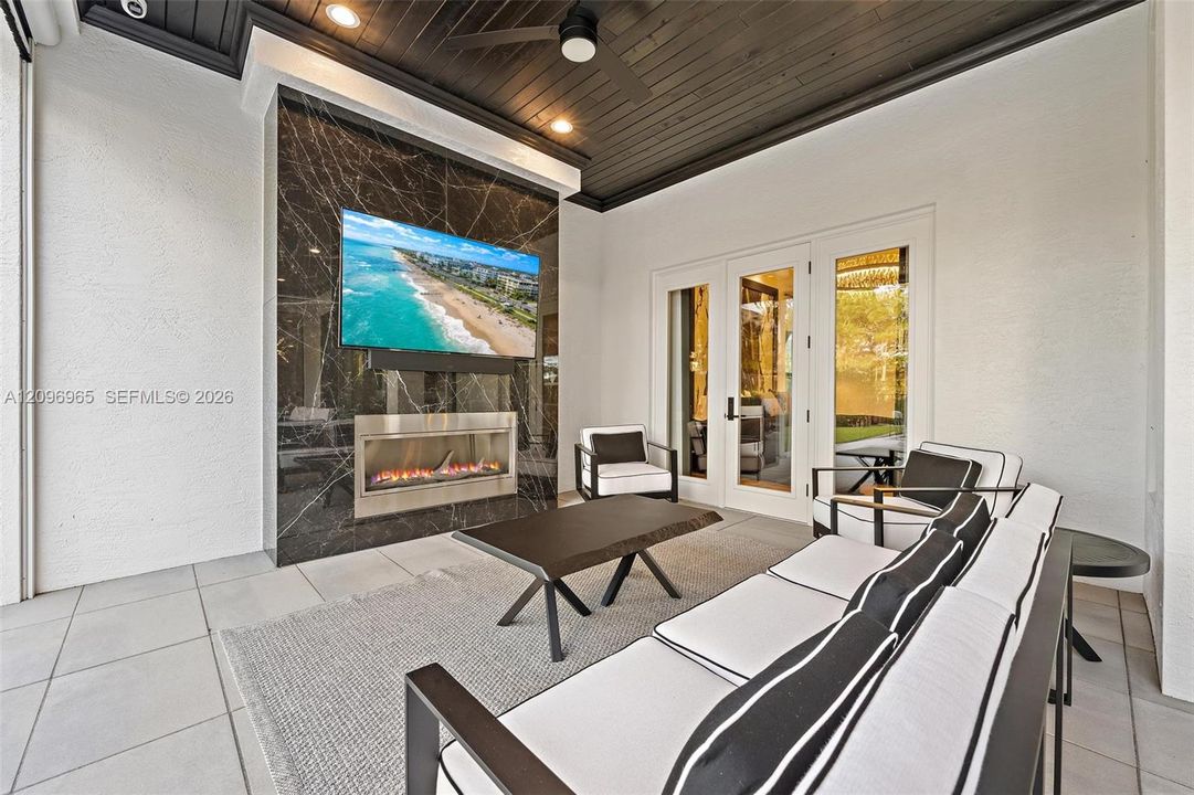 Covered patio featuring a floor-to-ceiling stone-cladding fireplace surround, stainless steel linear fireplace, black tongue-and-groove ceiling with integrated recessed lighting, ceiling fan, and large-format tile flooring