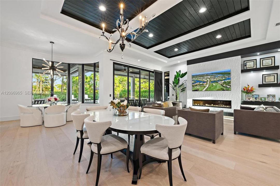 Coffered ceiling with black shiplap paneling and recessed lighting, wood-finish flooring, floor-to-ceiling glass panel walls, textured stone-surround linear fireplace, and integrated custom display shelving
