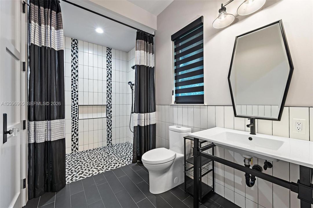 Bathroom featuring a vanity with a white countertop and matte black metal base, geometric wall mirror, vertical tile backsplash, and walk-in shower with decorative tile accent strips and a recessed niche
