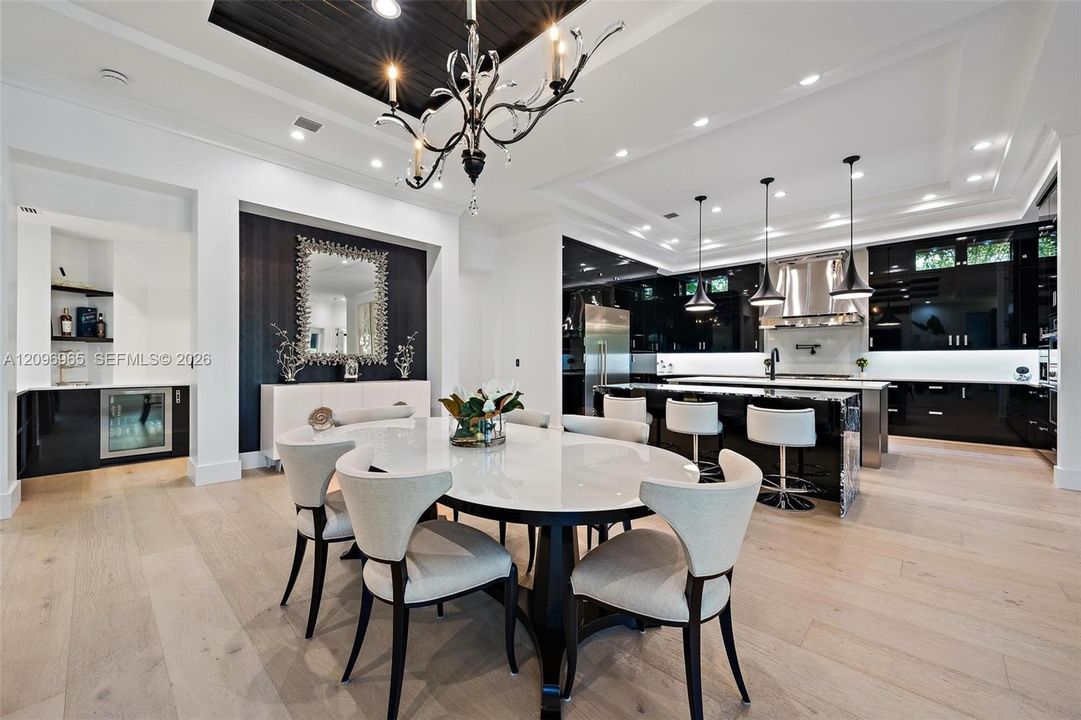 Open-concept kitchen and dining area featuring light wood-finish flooring, high-gloss cabinetry, a coffered ceiling, a recessed tray ceiling with dark wood inlay, and a built-in beverage refrigerator