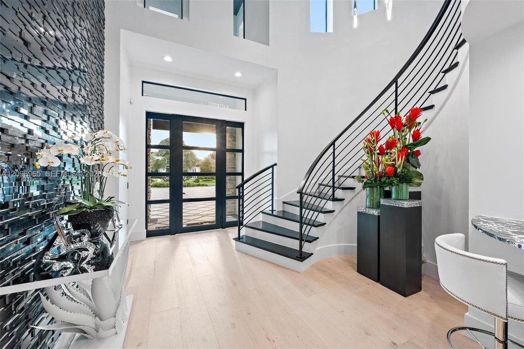 Grand entryway featuring high ceilings, a custom curved staircase with matte black metal railings, light-toned wood-finish flooring, a multi-pane glass-panel entry door, and a textured decorative accent wall