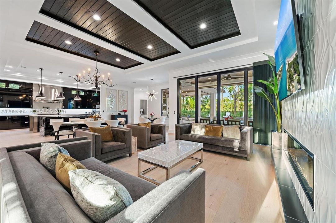 Coffered ceilings with dark-stained wood accents, light-toned wood-finish flooring, recessed lighting, expansive multi-panel glass doors, and a textured-tiled wall with a built-in fireplace