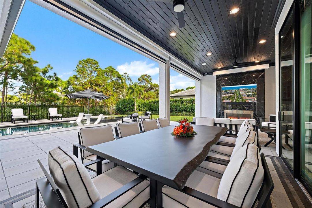 Covered outdoor living space featuring a dark-stained wood-plank ceiling, recessed lighting, integrated ceiling fans, and a built-in fireplace with tile surround, extending to a tiled patio and pool area