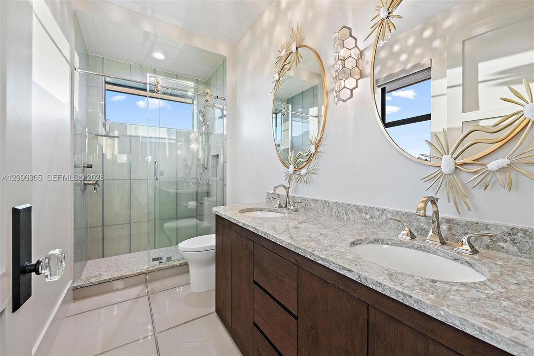 Dual-sink vanity with stone-finish countertop, dark wood-tone cabinetry, glass-enclosed shower featuring vertical tile, and large-format porcelain floor tiling