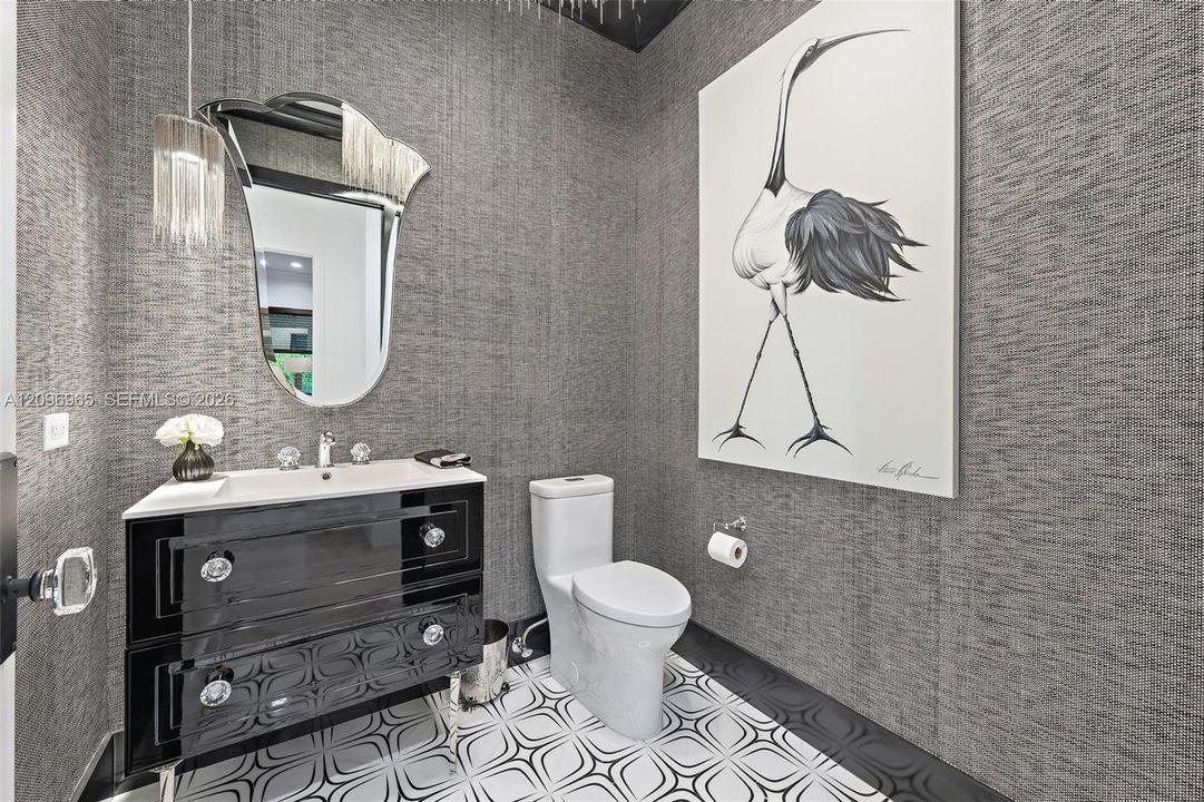 Textured dark grey wall coverings, black high-gloss cabinetry with crystal-style hardware, white vanity top with chrome faucet, geometric pattern tile flooring, and a decorative wall-mounted mirror