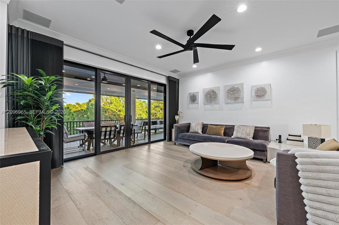 Living area featuring light-toned wood-finish flooring, multi-panel sliding glass doors with black frames, a matte black ceiling fan, and recessed ceiling lighting