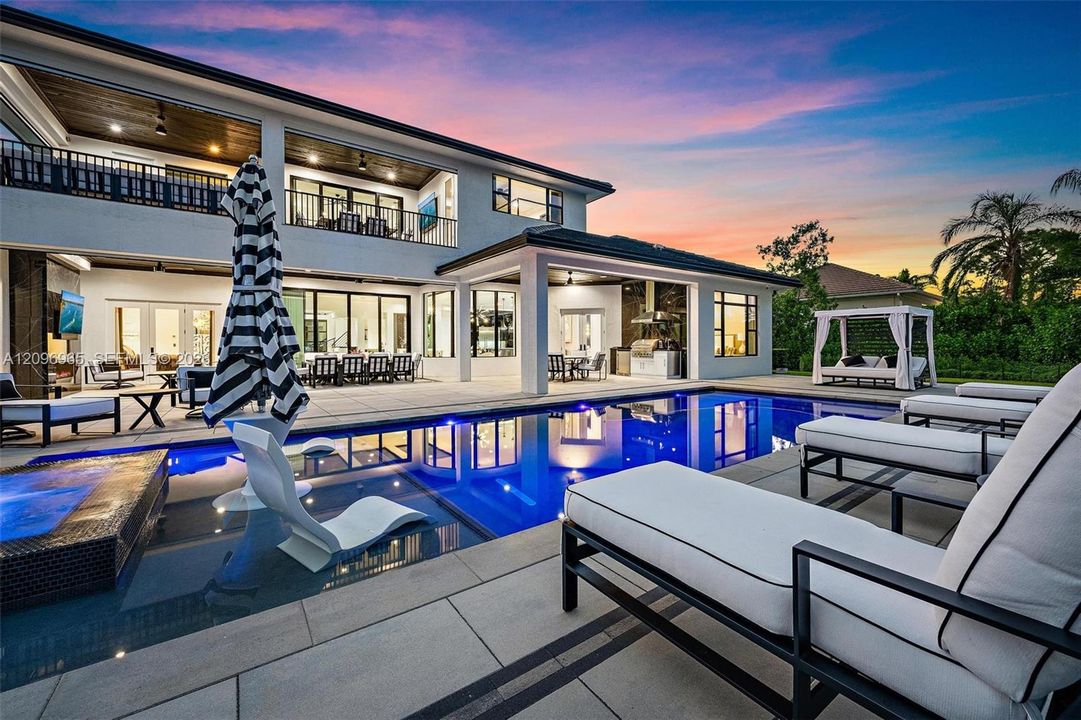 Multi-level exterior featuring a covered patio, built-in outdoor kitchen, rectangular swimming pool with integrated spa, and expansive stone-paved pool deck