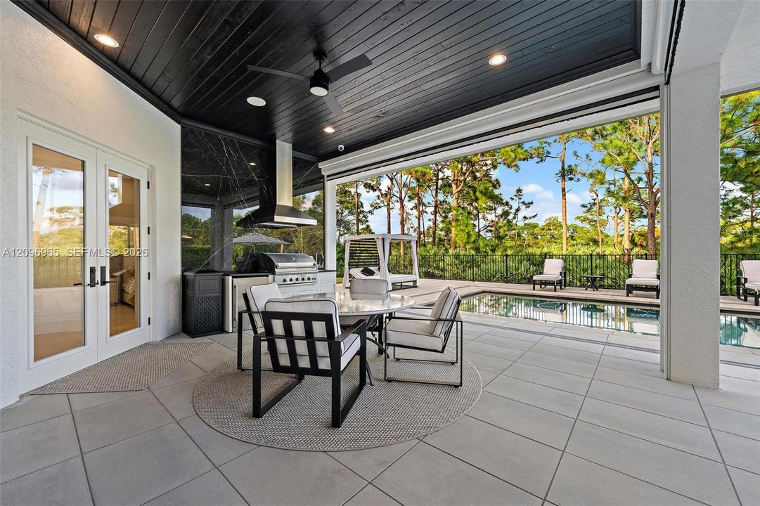 Covered outdoor kitchen featuring a stainless steel grill, vent hood, large-format tile flooring, a dark-stained wood ceiling with recessed lighting, and a swimming pool