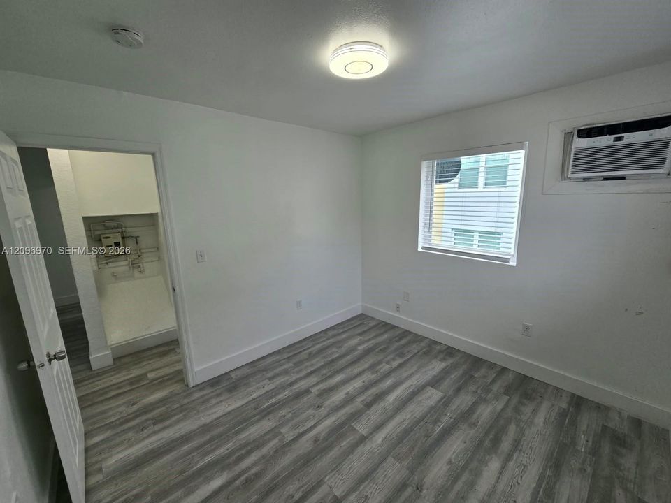 Neutral-toned room featuring wood-finish flooring, a single-hung window with installed blinds, a wall-mounted air conditioning unit, and a flush-mount ceiling light