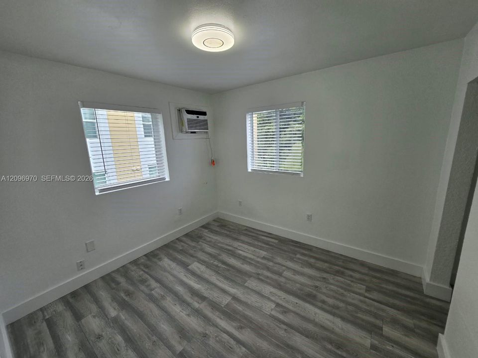 Neutral-toned wood-finish flooring, two windows with horizontal blinds, flush-mount ceiling light fixture, and wall-mounted air conditioning unit