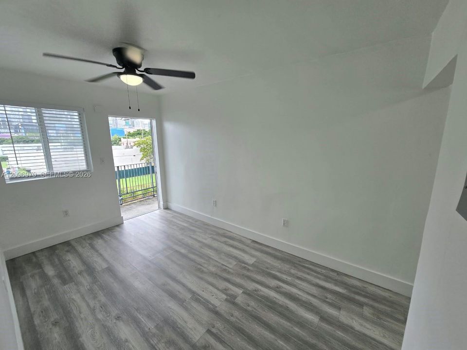 Interior space featuring wood-finish flooring, a multi-blade ceiling fan with integrated light fixture, a window with horizontal blinds, and access to a balcony with metal railing