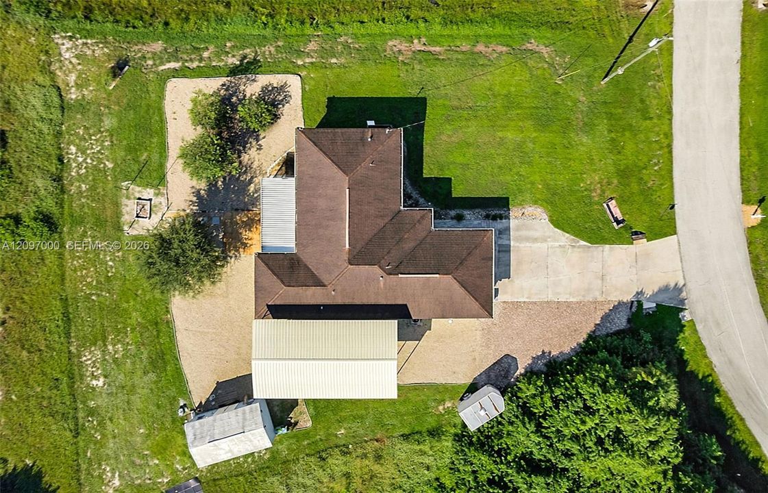 Aerial perspective of property featuring a shingled hipped roof, concrete driveway, metal-roofed detached structure, gravel lot, and landscaped grounds