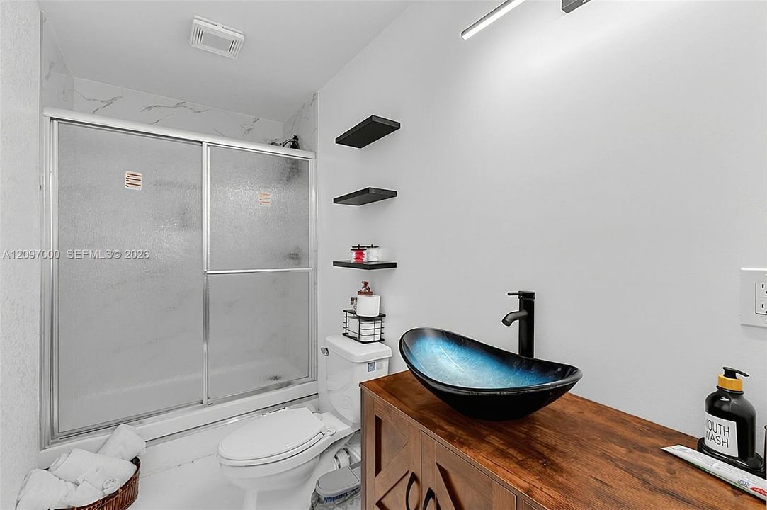 Bathroom featuring a wood-finish vanity with a vessel sink, matte black faucet, glass-enclosed shower, and wall-mounted shelving
