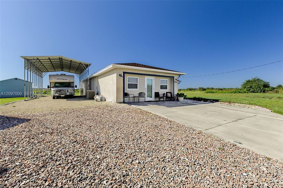 Stucco exterior, covered metal carport, expansive gravel driveway, and concrete patio