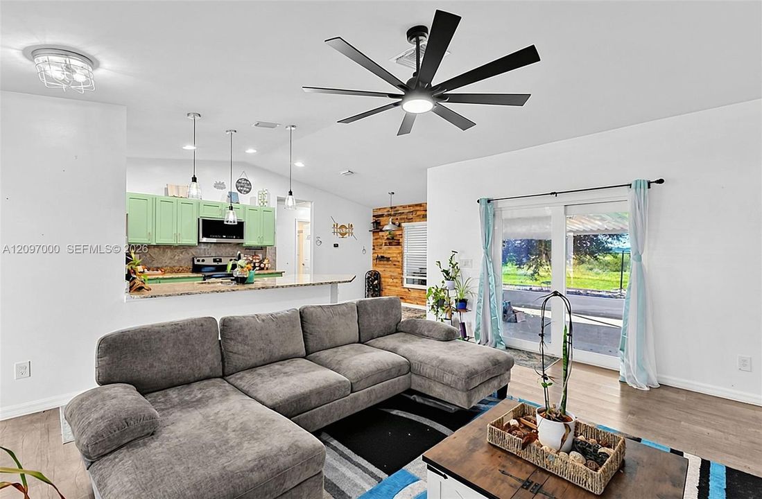 Open-concept interior featuring wood-finish flooring, vaulted ceiling with a multi-blade matte black fan, kitchen breakfast bar with stone-finish countertops, and dual sliding glass doors