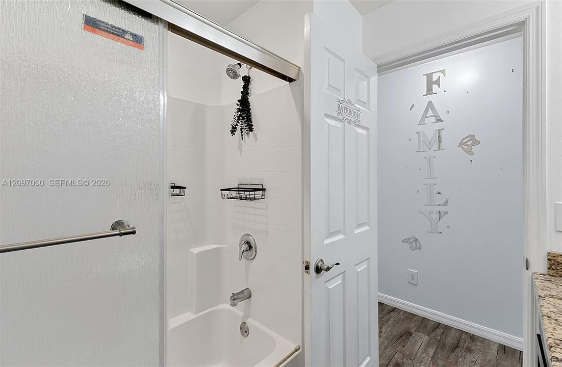 Tub and shower combination with a sliding glass door, white wall tile surround, brushed nickel hardware, and wood-finish flooring