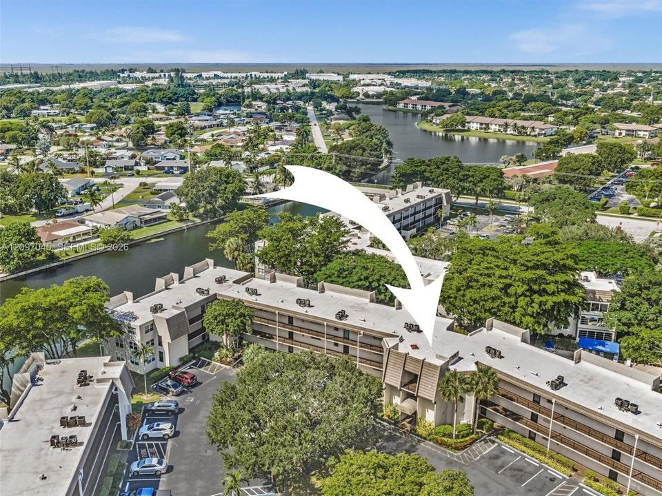 Aerial perspective featuring a multi-story building with a flat roof, textured exterior facade, designated surface parking, and extensive mature landscaping located along a canal