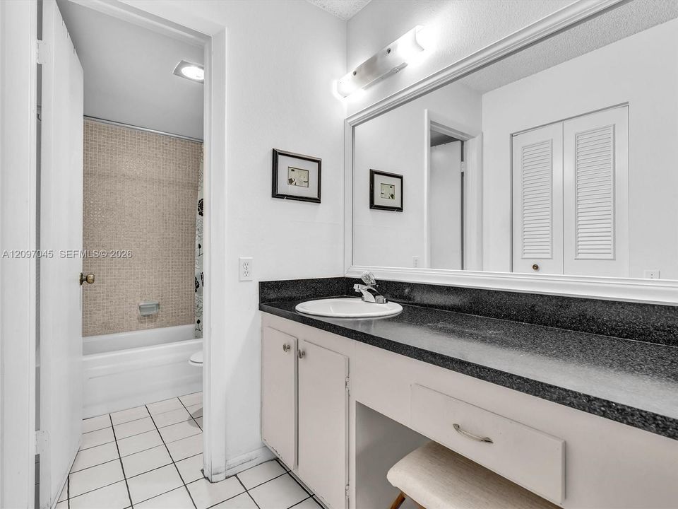 Vanity featuring a dark stone countertop, integrated circular basin, and full-width wall mirror with a mounted light fixture, complemented by tiled flooring and a shower-tub combination with mosaic wall tile