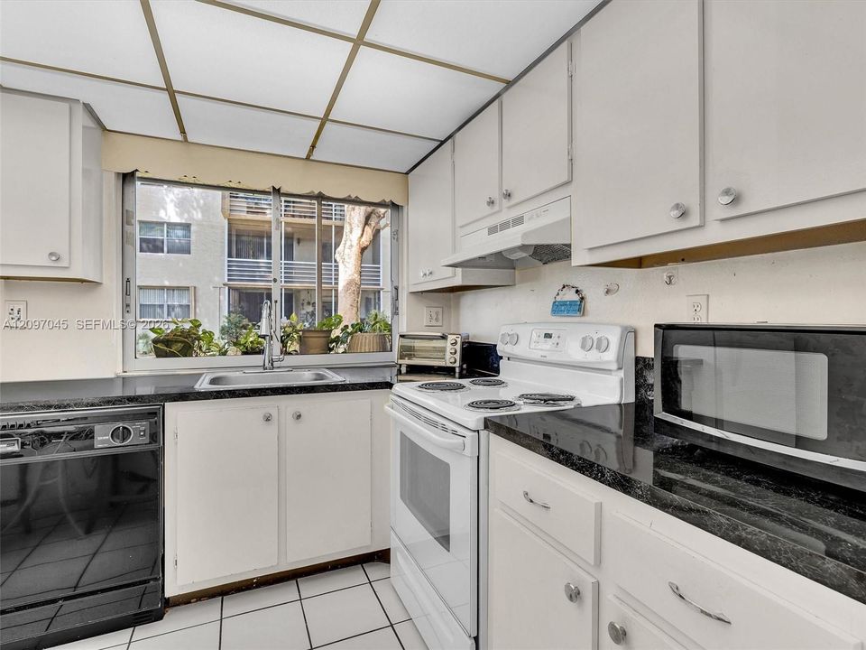 Kitchen featuring white cabinetry, dark stone countertops, an integrated range hood, white tile flooring, and a large window above the stainless steel sink
