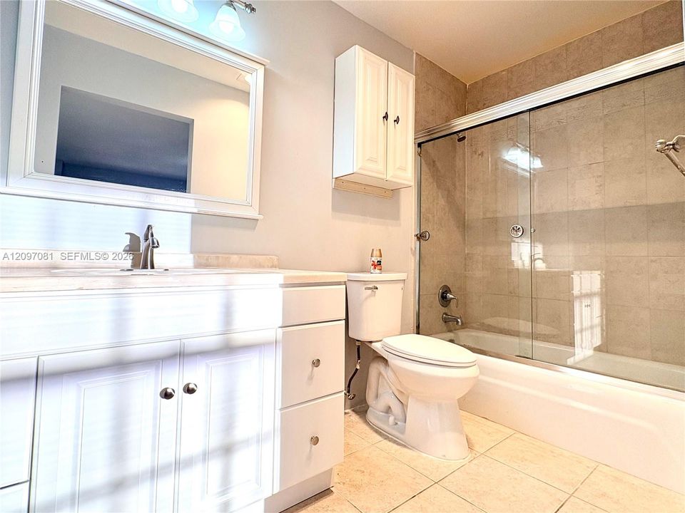 Bathroom featuring white shaker-style cabinetry, framed vanity mirror, wall-mounted storage cabinet, tile-surround tub with sliding glass doors, and light-toned floor tile