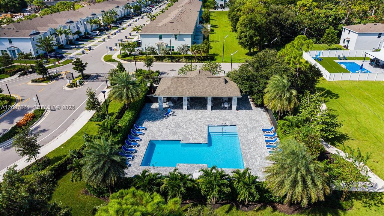 Community swimming pool with an L-shaped basin, expansive paved pool deck, covered pavilion, and mature landscaping
