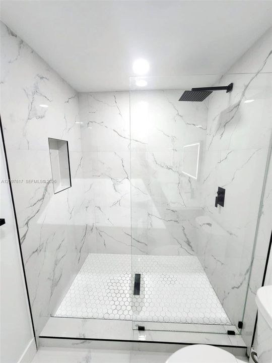 Walk-in shower featuring large-format white stone-finish wall tile, hexagonal floor tiling, a frameless glass enclosure, a built-in recessed niche, and matte black plumbing fixtures