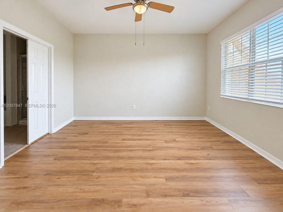 Neutral-toned interior featuring wood-finish flooring, a multi-blade ceiling fan with integrated lighting, and a window fitted with horizontal blinds
