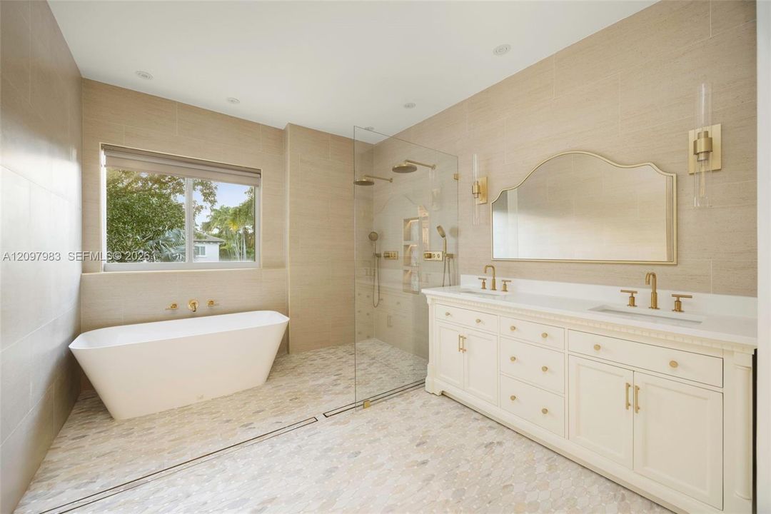 Freestanding soaking tub, glass-enclosed shower with dual rainfall showerheads, custom white cabinetry with integrated dual sinks, brass-finish hardware and fixtures, and textured wall tile