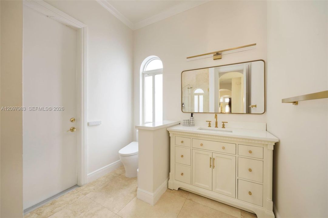 Bathroom featuring a white vanity with gold-tone hardware, stone-finish tile flooring, an arched window, and integrated vanity lighting