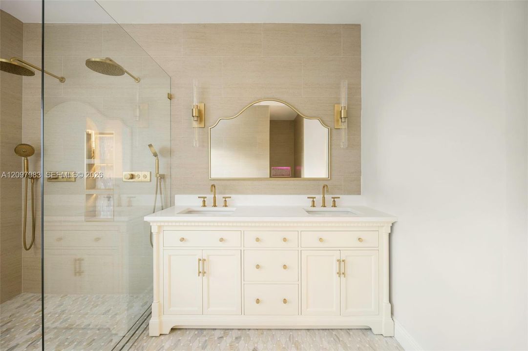 Double vanity with a white countertop and gold-toned hardware, arched wall mirror, wall-mounted brass light fixtures, and a glass-enclosed shower featuring dual rainfall showerheads and horizontal tile wall surrounds