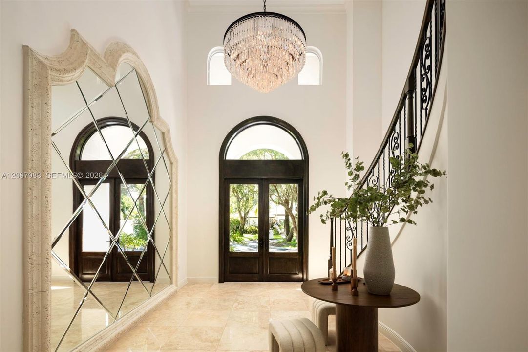Foyer featuring a grand crystal chandelier, arched double entry doors, a large-scale decorative mirror with a stone-finish frame, stone flooring, and a curved staircase with ornate iron railing