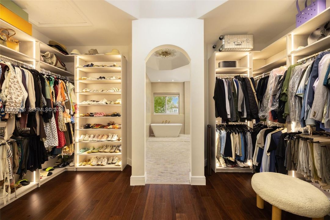 Walk-in closet featuring extensive custom built-in shelving, integrated vertical LED lighting, wood-finish flooring, and a central arched entry leading to a soaking tub