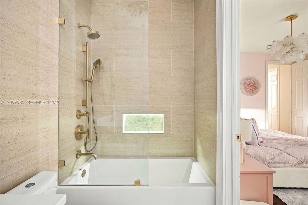 Bathtub with a glass panel, brass-finish rainfall showerhead and handheld sprayer, stone-tile wall surround, and a recessed tiled niche