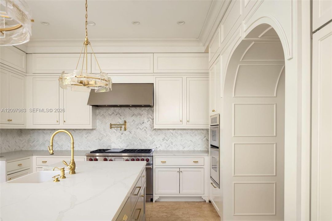Kitchen featuring custom cabinetry, herringbone-pattern stone tile backsplash, brass-finish fixtures, quartz countertops, and a paneled arched entryway