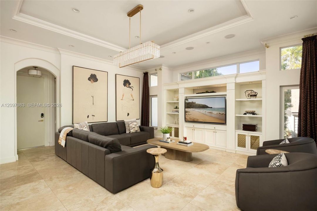 Living area featuring high ceilings with crown molding, custom built-in cabinetry, large-format tile flooring, arched entryway, and a tiered linear chandelier