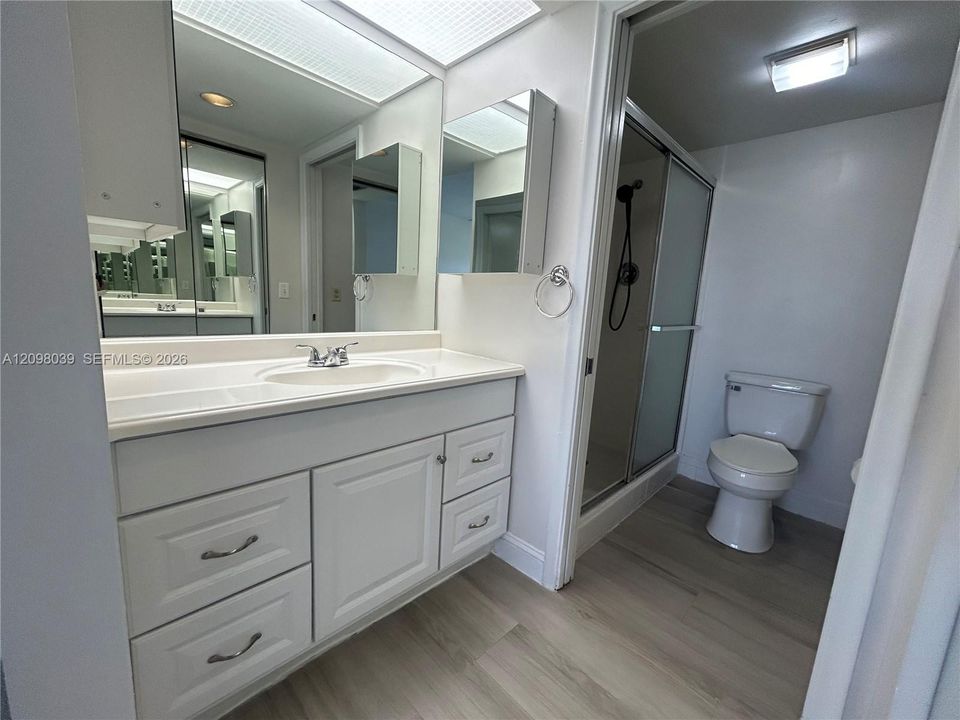 Bathroom featuring a white vanity with integrated basin, full-width wall mirror, wood-finish flooring, and a glass-enclosed shower